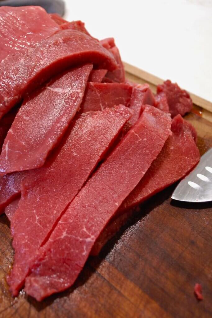Thinly sliced rump roast cut against the grain on a wooden cutting board for making tender beef jerky.